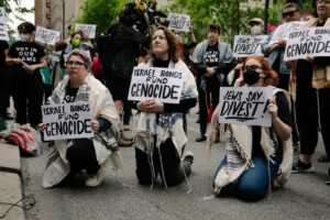 A group of people kneel while holding signs that say "Israel Bonds Fund Genocide."
